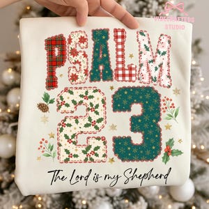 May include: Cream-colored t-shirt featuring the text "PSALM 23" in a patchwork design with plaid, holly, and floral patterns. The number "23" is prominently displayed, and the phrase "The Lord is my Shepherd" is written below.