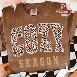May include: A brown Comfort Colors t-shirt with the words "COZY SEASON" in a combination of leopard print and tan block letters. The shirt has a crew neck and short sleeves. The brand name "VIBECRAFTED STUDIO" is printed on the upper right.