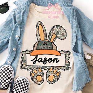 May include: A cream-colored t-shirt featuring a cartoon bunny wearing a hat that says "Egg Hunter" and the name "Jason". The bunny has camouflage ears and paws. The shirt is paired with a denim jacket and checkered shoes.