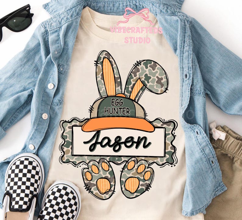May include: A cream-colored t-shirt featuring a cartoon bunny wearing a hat that says "Egg Hunter" and the name "Jason". The bunny has camouflage ears and paws. The shirt is paired with a denim jacket and checkered shoes.