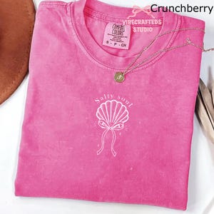 May include: A hot pink t-shirt with a white seashell graphic and the words "Salty soul". The shirt has a crew neck and short sleeves. A gold necklace with a small pendant is draped across the shirt.