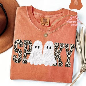 May include: A burnt orange short-sleeved t-shirt featuring the word SPOOKY in a varsity-style font. The letters S, P, K, and Y are filled with a brown and black leopard print, while the two O's are replaced by white lace ghost figures.