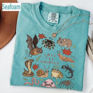 May include: Seafoam green t-shirt featuring a graphic of a bat, spider, mouse, and snail. The text "LOVE THE UNLOVED" is printed below the illustrations. The shirt has a crew neck and short sleeves. The label reads "Comfort Colors".