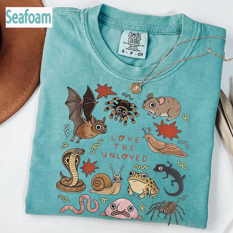 May include: Seafoam green t-shirt featuring a graphic of a bat, spider, mouse, and snail. The text "LOVE THE UNLOVED" is printed below the illustrations. The shirt has a crew neck and short sleeves. The label reads "Comfort Colors".