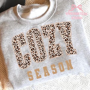 May include: A gray sweatshirt with the words "COZY SEASON" printed on the front. The word "COZY" is in a leopard print pattern, and the word "SEASON" is in a tan block letter font.