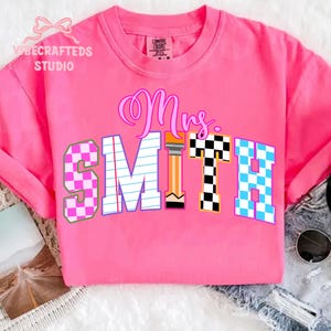 May include: A bright pink t-shirt with the text "Mrs. SMITH" in a colorful, school-themed design. The "S" and "H" are checkered, the "M" is lined like notebook paper, and the "T" is a pencil.