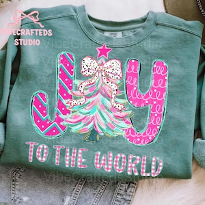 May include: A teal green sweatshirt with a colorful Christmas tree design and the words "JOY TO THE WORLD." The tree is pink and teal with a polka dot bow. The letters are pink with patterns.