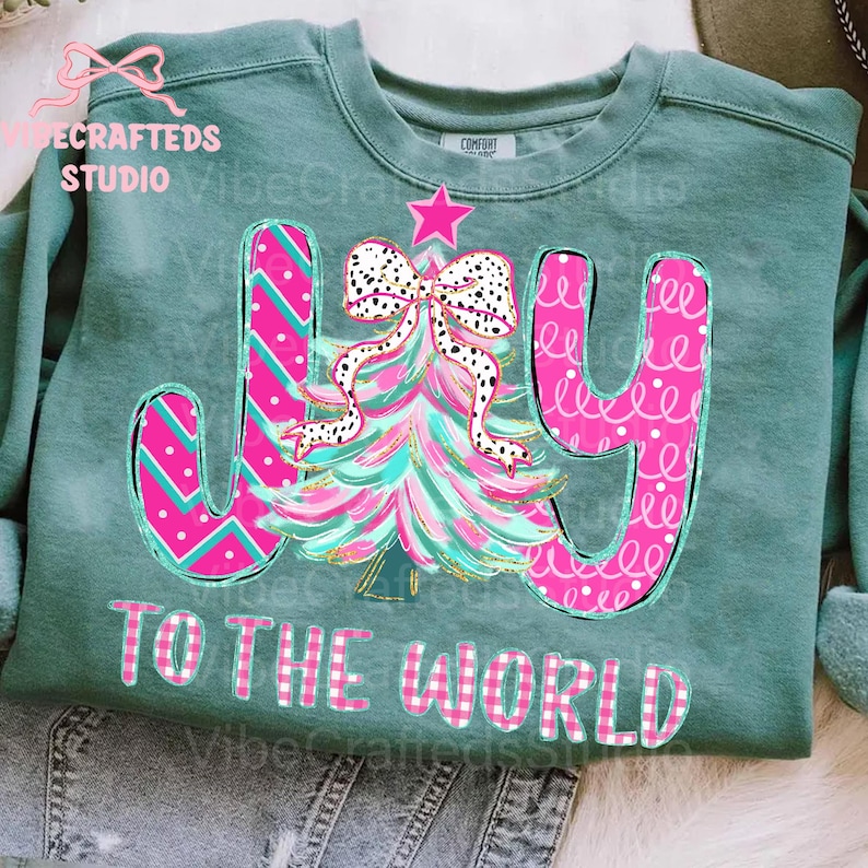 May include: A teal green sweatshirt with a colorful Christmas tree design and the words "JOY TO THE WORLD." The tree is pink and teal with a polka dot bow. The letters are pink with patterns.