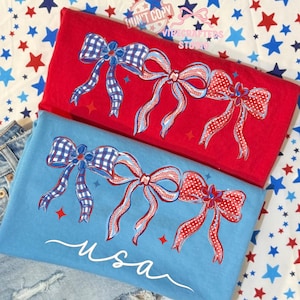 May include: Red and blue t-shirts with bow designs. One shirt is red, the other is blue, and both feature a bow design with red, white, and blue accents. The blue shirt has the word "usa" in white script.