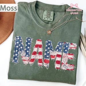 May include: A moss green t-shirt with the word "NAME" appliqued across the front in a patriotic design. The letters feature a patchwork of red and white stripes and blue with white stars, resembling the American flag. A silver necklace with a pendant is also visible.
