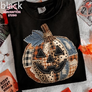 Könnte beinhalten: Ein schwarzes T-Shirt mit einem großen, zentralen Kürbis-Applikationsmotiv aus verschiedenen Stoffstücken wie Denim, Karomuster, Leopardenmuster und Spitze. Der Kürbis hat ein aufgenähtes Gesicht aus schwarzem Stoff und ein ausgefranstes Denim-Blatt.