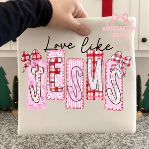 May include: Cream-colored tote bag with the words "Love like JESUS" in black script. The word JESUS is in pink and red with heart and bow accents. The bag is held by a hand.