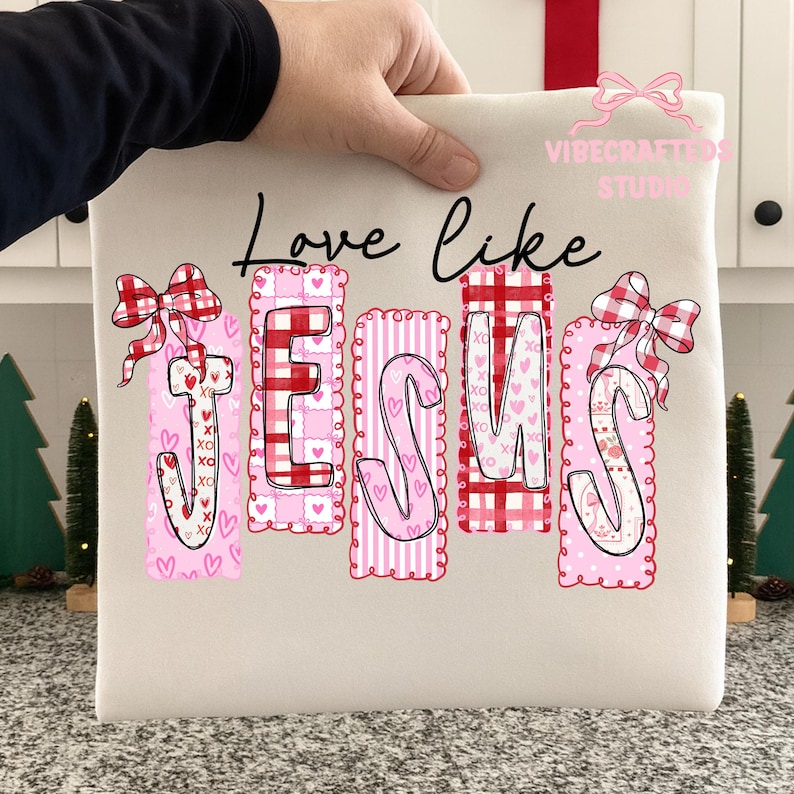 May include: Cream-colored tote bag with the words "Love like JESUS" in black script. The word JESUS is in pink and red with heart and bow accents. The bag is held by a hand.