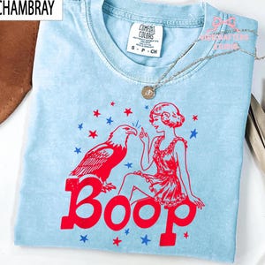 May include: Pale blue t-shirt featuring a red graphic of an eagle, a woman, and the word "Boop." The design is surrounded by red and blue stars. The shirt has a crew neck and short sleeves.