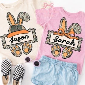 May include: Two short-sleeved t-shirts, one cream and one pink, feature a bunny design with camouflage ears and paws. The cream shirt has the name "Jason" on a banner, and the pink shirt has "Sarah". A pair of checkered slip-on shoes and blue shorts are also visible.
