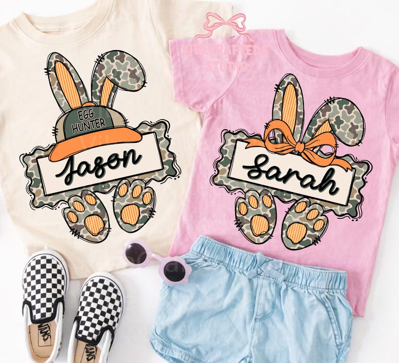 May include: Two short-sleeved t-shirts, one cream and one pink, feature a bunny design with camouflage ears and paws. The cream shirt has the name "Jason" on a banner, and the pink shirt has "Sarah". A pair of checkered slip-on shoes and blue shorts are also visible.