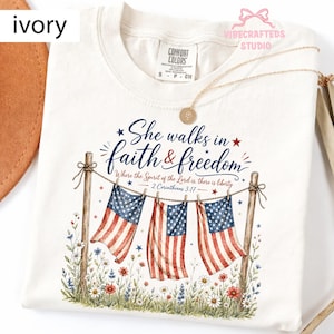 Könnte beinhalten: Elfenbeinfarbenes T-Shirt mit dem Text "She walks in faith & freedom" und amerikanischen Flaggen-Grafiken. Das Shirt hat einen Rundhalsausschnitt und kurze Ärmel. Das Design umfasst florale Akzente und den Bibelvers 2. Korinther 3:17.