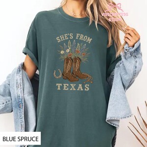 May include: Blue spruce t-shirt with the text "She's From Texas" and a graphic of brown cowboy boots, flowers, and a horseshoe. The shirt has short sleeves and a relaxed fit.
