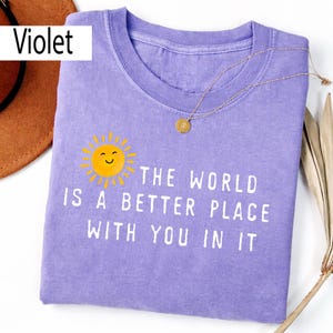 May include: A violet crew neck t-shirt featuring a yellow sun graphic with a smiling face and the white text "THE WORLD IS A BETTER PLACE WITH YOU IN IT". The shirt has a short-sleeved design and a ribbed collar, presented in a flat lay view.