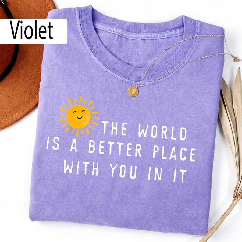 May include: A violet crew neck t-shirt featuring a yellow sun graphic with a smiling face and the white text "THE WORLD IS A BETTER PLACE WITH YOU IN IT". The shirt has a short-sleeved design and a ribbed collar, presented in a flat lay view.