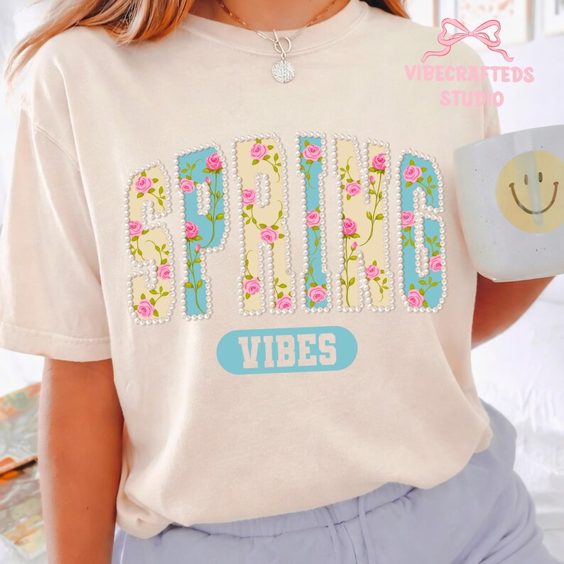 May include: Cream-colored t-shirt with the words "SPRING VIBES" in a floral design. The letters are outlined in light blue and adorned with pink roses and pearls. The word "VIBES" is in a light blue rectangle.
