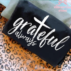 May include: Black t-shirt with the words "grateful always" and a white cross design. The shirt is folded and rests on a leopard print fabric. Additional fabric in shades of green and blue are visible in the background.