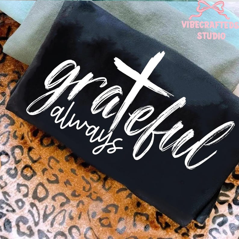 May include: Black t-shirt with the words "grateful always" and a white cross design. The shirt is folded and rests on a leopard print fabric. Additional fabric in shades of green and blue are visible in the background.