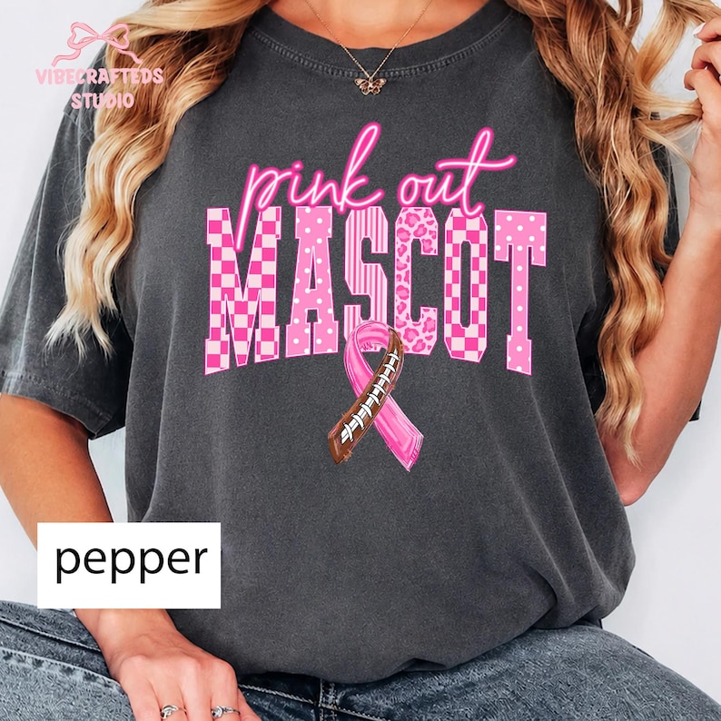 May include: A charcoal gray short-sleeve t-shirt featuring the pink text "pink out" above the word "MASCOT" in a block font. The letters contain various pink patterns including checkers, stripes, and leopard print, with a football-themed ribbon.