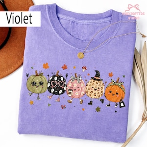 Pode incluir: Uma t-shirt violeta de manga curta com uma impressão gráfica de cinco abóboras antropomórficas com vários acessórios, incluindo óculos em forma de estrela, óculos de coração, um laço, um chapéu de bruxa e uma fita, rodeadas por folhas de outono.