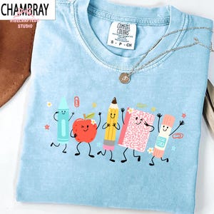 Puede incluir: Una camiseta de cuello redondo azul claro con un estampado gr&aacute;fico de cinco &uacute;tiles escolares antropom&oacute;rficos, incluyendo un cray&oacute;n azul, una manzana roja, un l&aacute;piz amarillo, un cuaderno rosa y un pegamento en barra, todos con caras sonrientes.