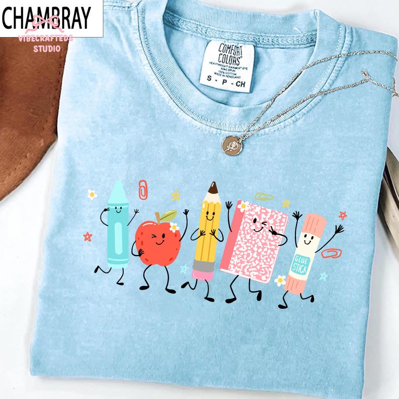 Puede incluir: Una camiseta de cuello redondo azul claro con un estampado gr&aacute;fico de cinco &uacute;tiles escolares antropom&oacute;rficos, incluyendo un cray&oacute;n azul, una manzana roja, un l&aacute;piz amarillo, un cuaderno rosa y un pegamento en barra, todos con caras sonrientes.