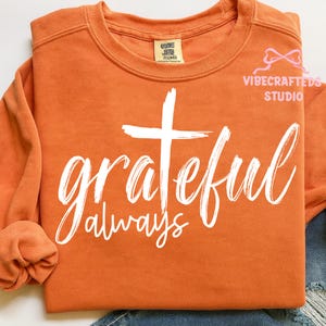 May include: Orange crewneck sweatshirt with the words "grateful always" in white script, and a white cross. The sweatshirt has long sleeves and a tag that says "comfort colors". The VIBECRAFTED STUDIO logo is in the upper right corner.