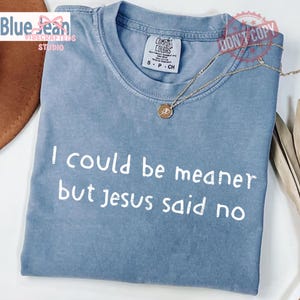 May include: A light blue t-shirt with the text "I could be meaner but Jesus said no" in white. The shirt has a crew neck and short sleeves. A gold necklace with a pendant rests on the shirt.