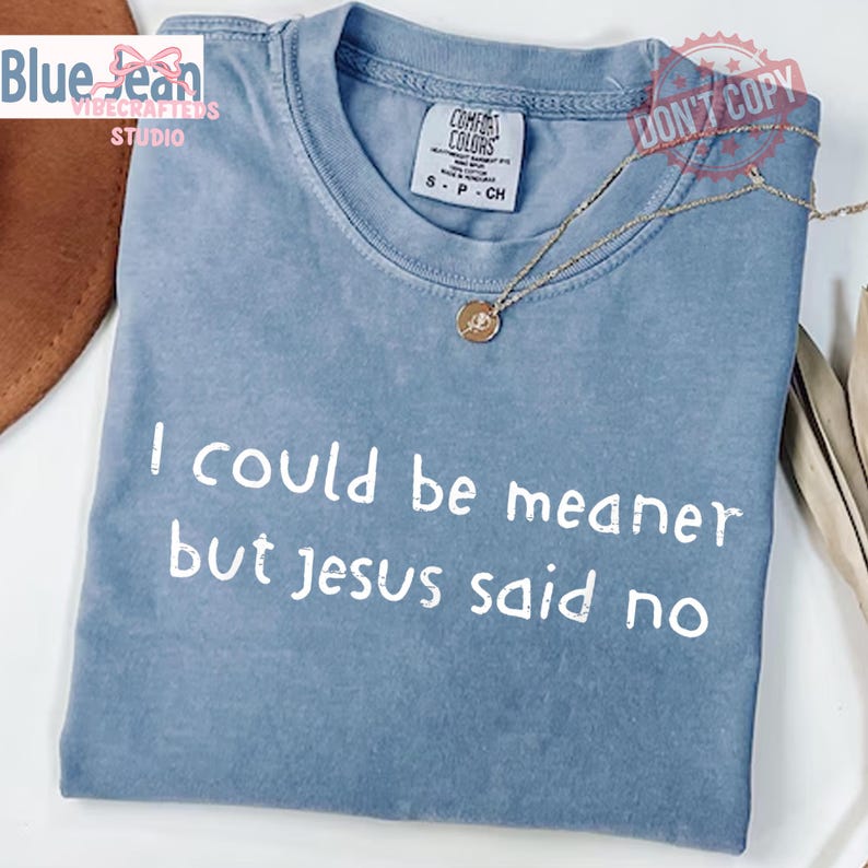 May include: A light blue t-shirt with the text "I could be meaner but Jesus said no" in white. The shirt has a crew neck and short sleeves. A gold necklace with a pendant rests on the shirt.
