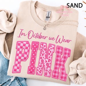 May include: A sand-colored crewneck sweatshirt featuring the text In October we Wear PINK in a script and block font. The letters are filled with various pink patterns including checkers, polka dots, leopard print with ribbons, and plaid.