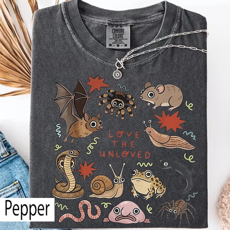 May include: Dark grey t-shirt featuring a collection of illustrated creatures such as a bat, spider, and mouse, with the text "LOVE THE UNLOVED". A silver necklace is draped over the shirt. The shirt is labelled "Pepper".