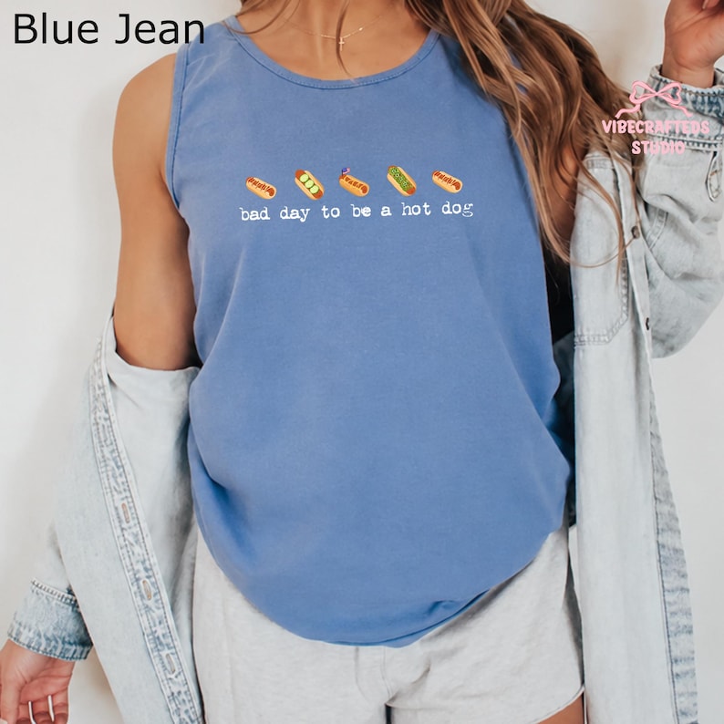 May include: A blue jean colored tank top featuring a graphic of five small hot dogs in a row above the white text that reads, bad day to be a hot dog. The sleeveless shirt has a round neckline and is worn with a light blue denim button-down shirt.