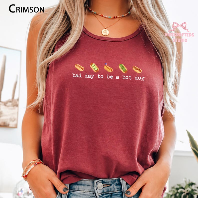 May include: A crimson sleeveless tank top featuring a graphic of five small hot dogs in a row above the white text "bad day to be a hot dog". The garment has a round neckline and is worn with a multi-colored beaded necklace and stacked bracelets.