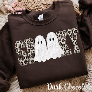 May include: A dark chocolate brown crewneck sweatshirt featuring the word SPOOKY in a leopard print pattern. The two letter O's are replaced by white lace ghost figures with black eyes. The sweatshirt has a ribbed collar and long sleeves.