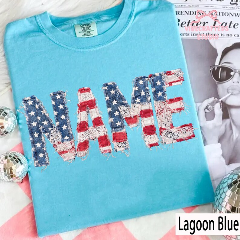 May include: A turquoise t-shirt featuring the word "NAME" in a patchwork design resembling the American flag. The shirt is labelled "Lagoon Blue" and has a "Current Colors" tag.