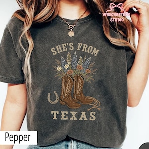 May include: Dark gray t-shirt with the text "She's From Texas" above a graphic of brown cowboy boots, flowers, and a horseshoe. The shirt is a Pepper color.