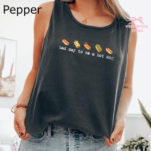 May include: A dark gray sleeveless tank top featuring a graphic of five hot dogs in various states of completion above the white text that reads, bad day to be a hot dog. The shirt has a round neckline and is worn with light blue denim jeans.