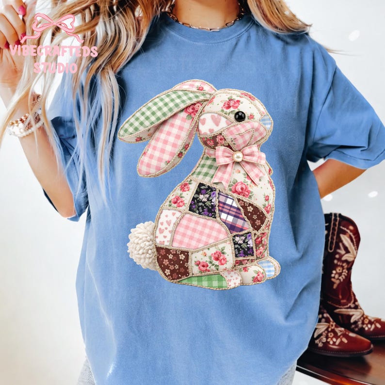 May include: A light blue t-shirt featuring a patchwork bunny design. The bunny is made of various patterned fabrics, including floral, plaid, and polka dot patterns. A pink bow adorns the bunny's neck. The shirt has the text "Vibe Crafted Studio" in the upper left corner.