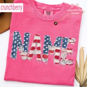 May include: A bright pink t-shirt featuring the word "NAME" in a patriotic design. The letters are crafted from denim with white stars and red and white striped fabric. The shirt also displays the text "crunchberry" and "Comfort Colors".