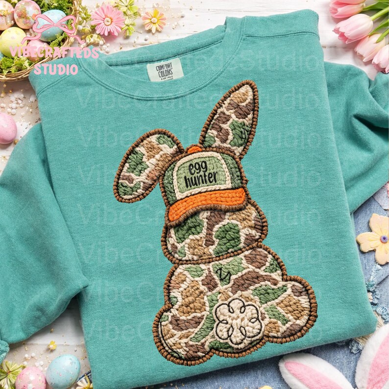 May include: A teal sweatshirt with a large embroidered bunny design. The bunny is in camouflage colors and wears a hat that says "Egg Hunter". The design is detailed with brown stitching.