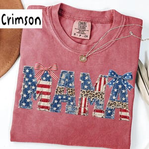 May include: Crimson t-shirt with the word "MAMA" in a patchwork design featuring American flag and leopard print patterns. The shirt has a Comfort Colors tag and a silver necklace with a pendant.