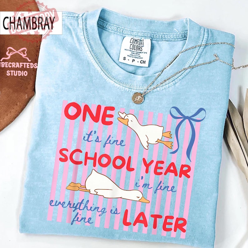 May include: Light blue t-shirt with the text "ONE it's fine SCHOOL YEAR i'm fine everything is fine LATER". The shirt features a graphic of two white geese and pink and blue accents. A silver necklace is draped across the shirt.