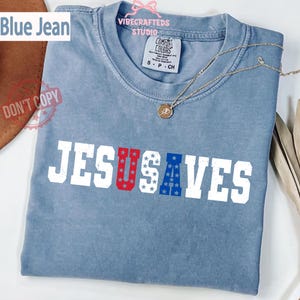 Könnte beinhalten: Ein blaues Jeans-T-Shirt mit den Worten "JESUS SAVES" auf der Vorderseite. Die Buchstaben "U" und "A" sind mit einem roten und blauen Sternmuster gefüllt. Eine goldene Halskette liegt auf dem Shirt.