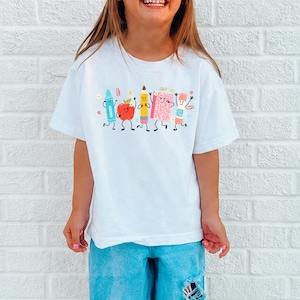 Puede incluir: Una camiseta blanca con un estampado gr&aacute;fico de cinco &uacute;tiles escolares antropom&oacute;rficos, incluyendo un cray&oacute;n azul, una manzana roja, un l&aacute;piz amarillo, un cuaderno rosa y un pegamento en barra, todos con caras sonrientes y extremidades negras.