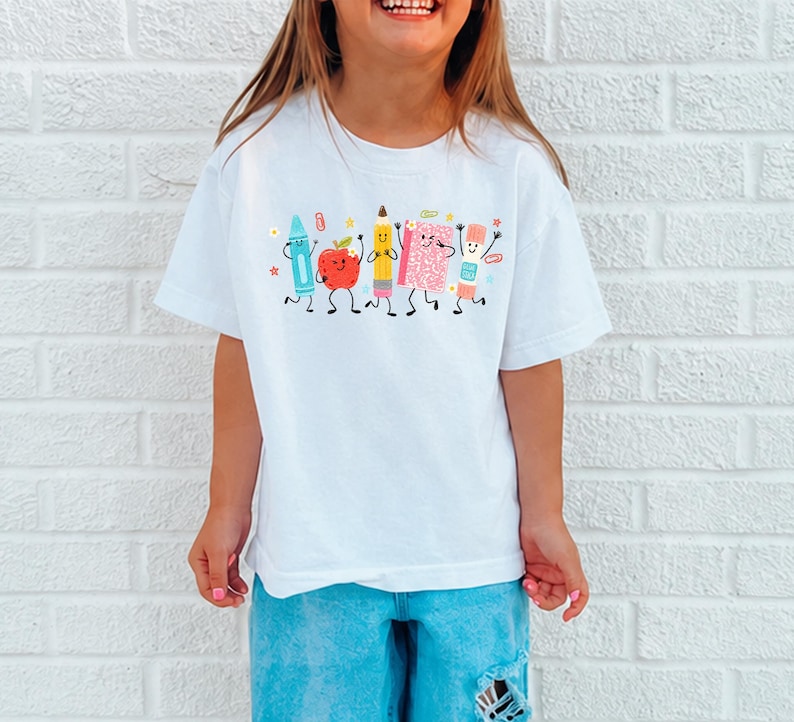 Puede incluir: Una camiseta blanca con un estampado gr&aacute;fico de cinco &uacute;tiles escolares antropom&oacute;rficos, incluyendo un cray&oacute;n azul, una manzana roja, un l&aacute;piz amarillo, un cuaderno rosa y un pegamento en barra, todos con caras sonrientes y extremidades negras.